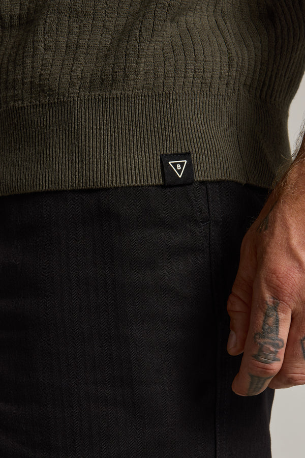 A close-up of a person wearing Barkers' Dawn Cotton Knit Tee in olive-green with ribbed stripes and black pants. A small black tag with a white triangular logo is visible, and their tattooed hand rests by their side.