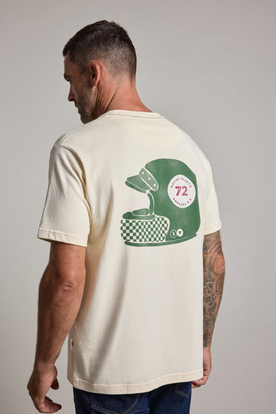 A man with short hair and a tattooed arm faces away, wearing the Barkers Vintage Helmet Print Tee in cream—a soft organic cotton shirt featuring a green helmet, checkered basket, and circular "72" badge graphic on the back.