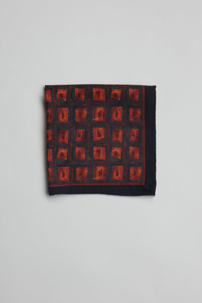 The Parisian Square Wool PS by Parisian is a folded dark blue 100% wool pocket square made in New Zealand, featuring a repeating red square and orange-red shape pattern, shown on a plain light gray background.