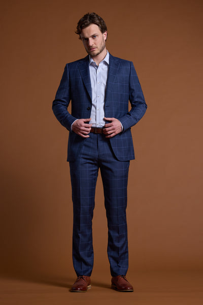 A man stands against a brown background wearing Barkers' Treyburn Check Suit Trousers, a white dress shirt, brown belt, and brown shoes. He buttons his jacket and looks at the camera in this temperature regulating suit.