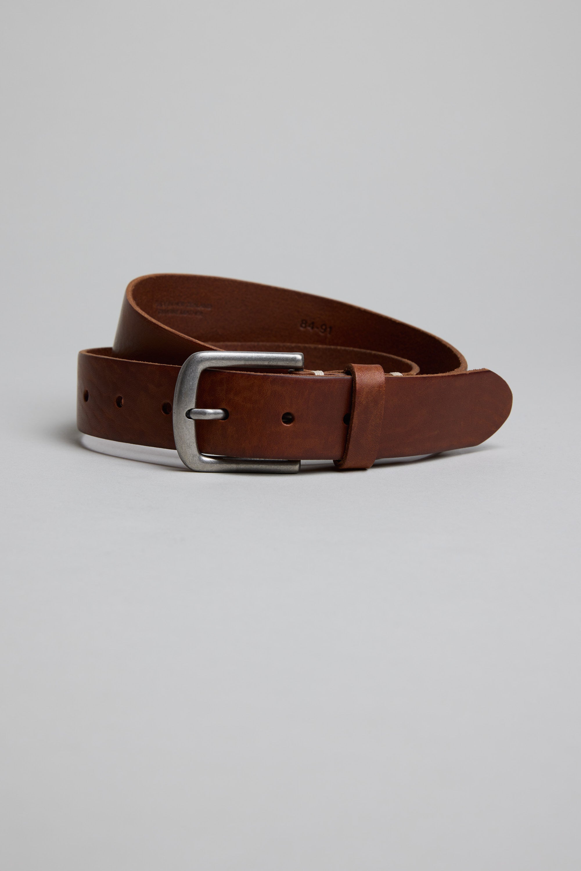 The Parisian Tuscany Leather Belt, made of genuine brown leather with a silver rectangular buckle, is shown coiled on a plain light grey background.