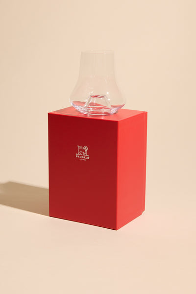 A clear, uniquely shaped Whisky Glass Individual by Other sits atop a bright red box with white branding, set against a beige background.