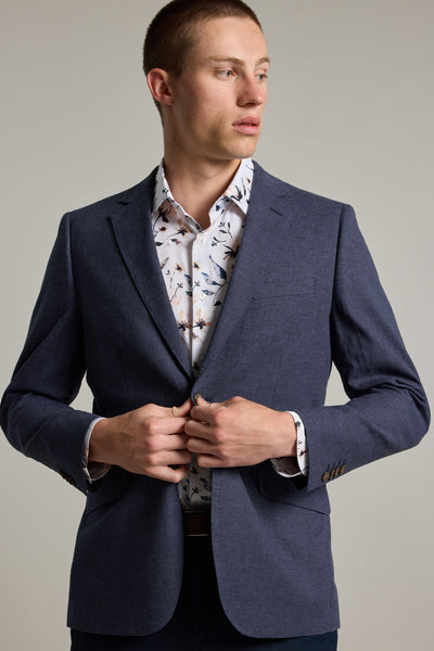 A man wearing the Barkers Ellwood Houndstooth Blazer over a white dress shirt with a bird pattern stands against a plain background, looking to his left while buttoning his jacket.
