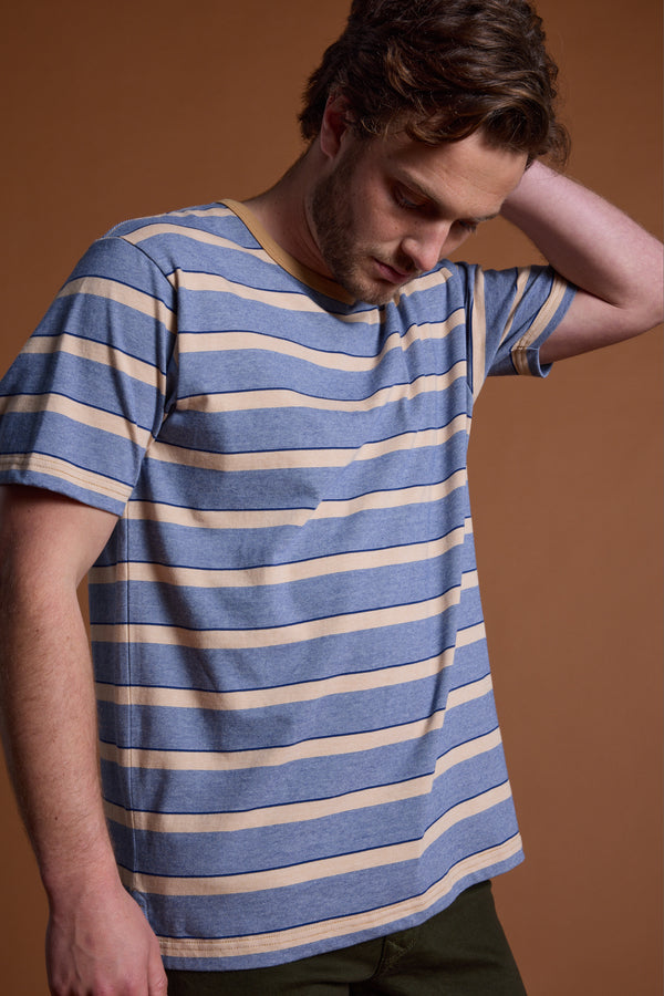 A man wearing the Barkers Chester Stripe Tee in blue and beige stands against a brown background, looking down with one hand on the back of his head.