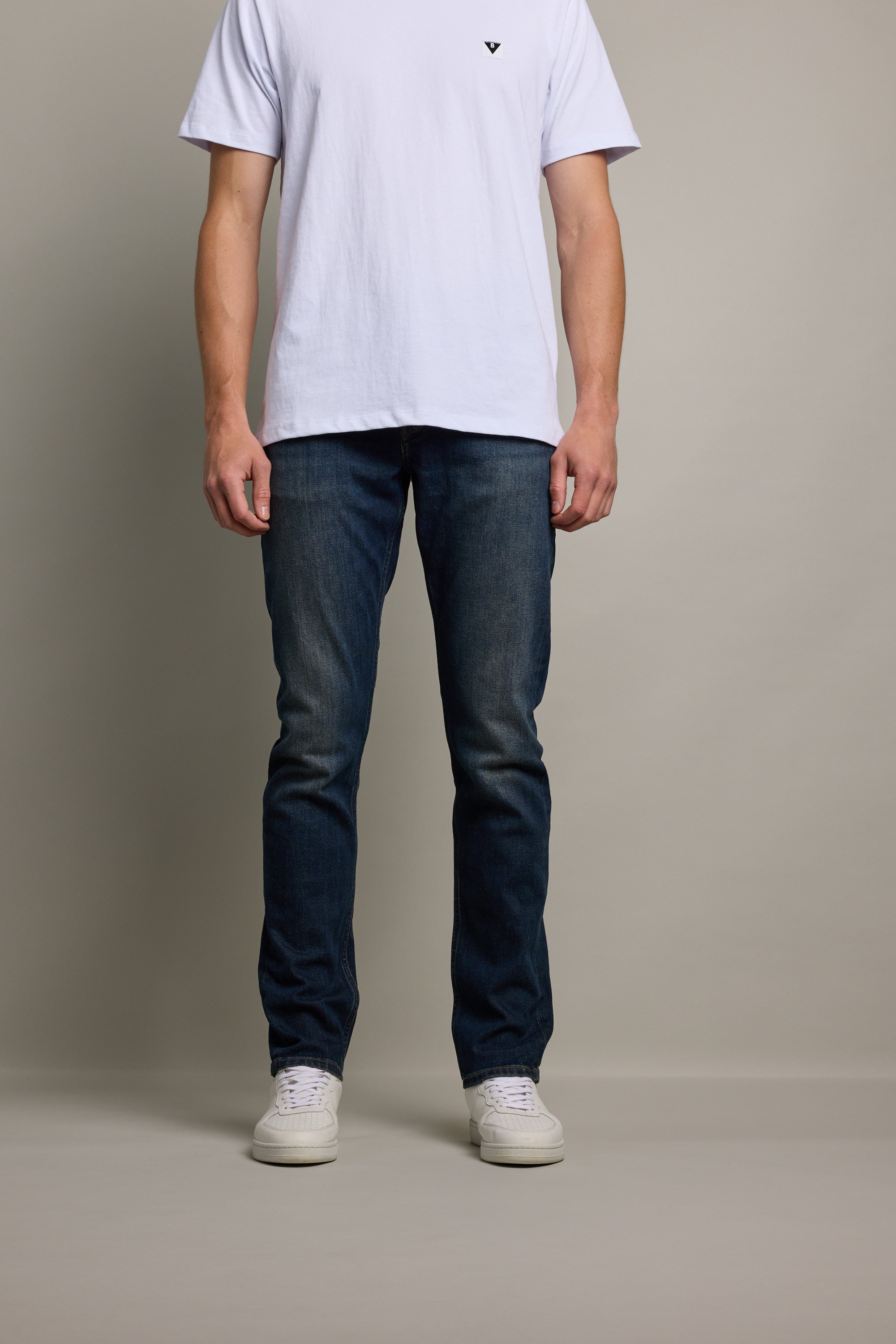 A person stands against a plain background, wearing a white short-sleeve t-shirt, Barkers Nomad Denim Taper Jean in dark blue, and white sneakers. The person's head is not visible in the image.