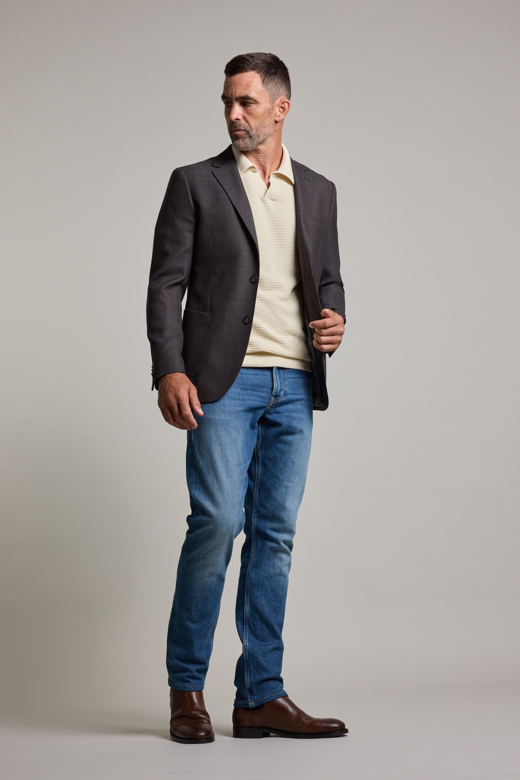 A man stands against a plain background wearing the Barkers Riverside Texture Blazer over a cream shirt, blue jeans, and brown boots. He looks slightly left, adjusts his blazer with one hand, and maintains a neutral expression.