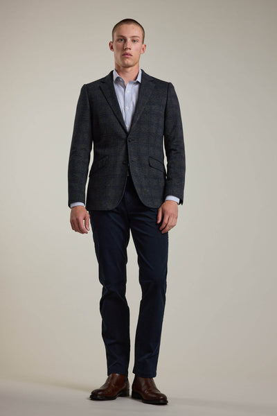 A man faces forward against a plain background, wearing the Barkers Manleigh Check Knit Blazer, a light blue dress shirt, dark blue trousers, and brown dress shoes with a neutral expression.