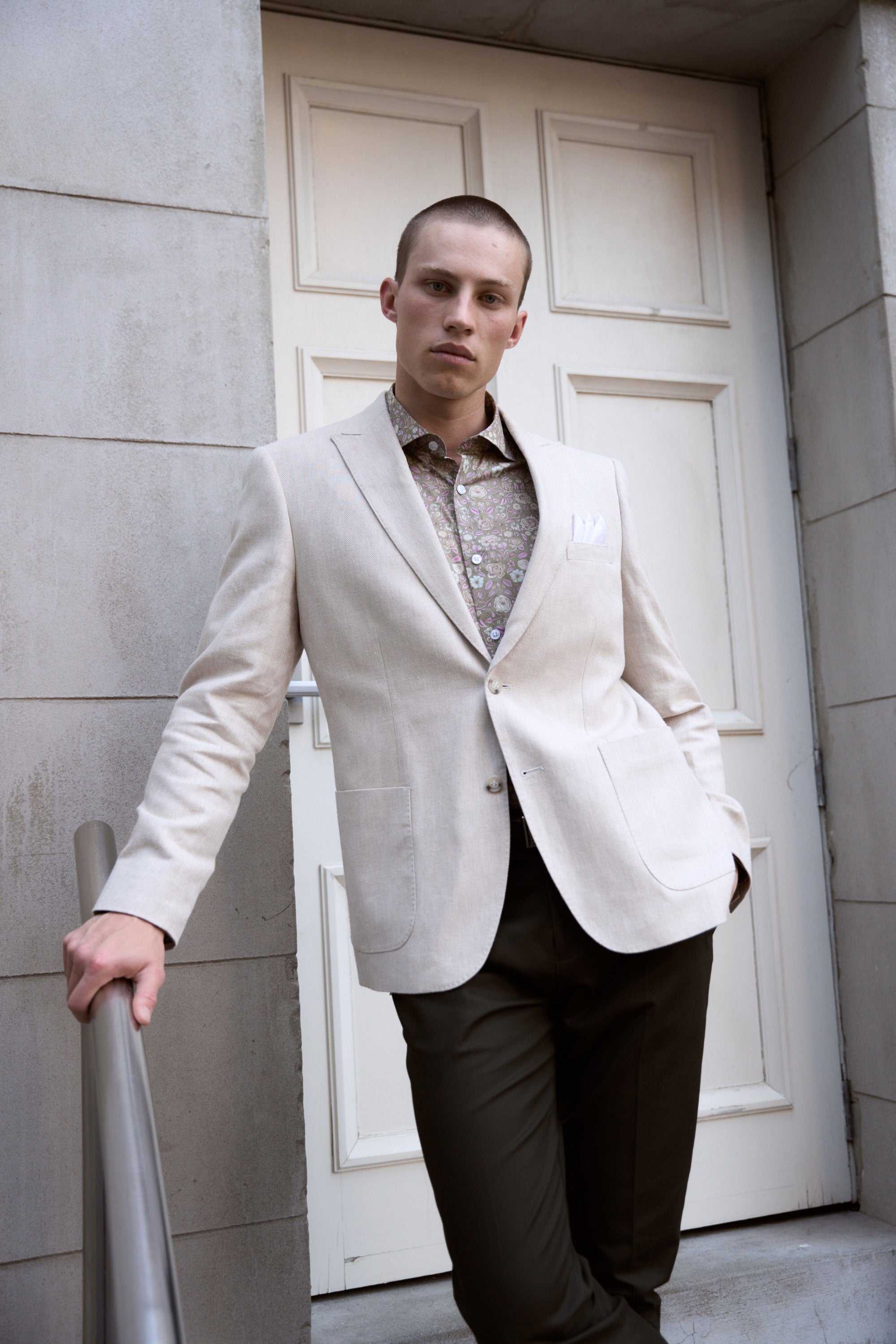 A man wearing the Barkers Hume Herringbone Blazer, a patterned shirt, and dark pants stands confidently by a metal railing before a white panel door—stylish summer event attire.