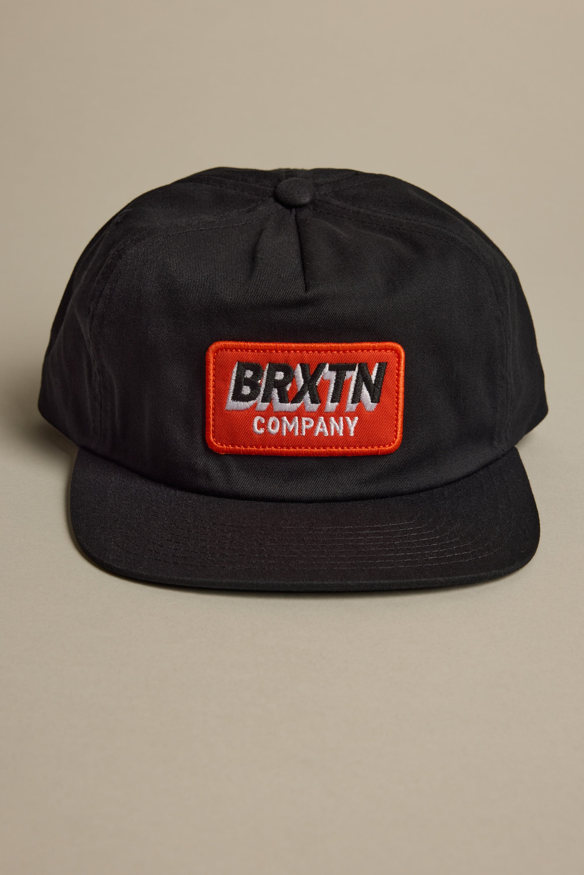 The Brixton Payment MP Snapback, a black 5-panel hat with a flat brim, features a red and white BRXTN COMPANY patch on the front. It is shown against a neutral beige background.