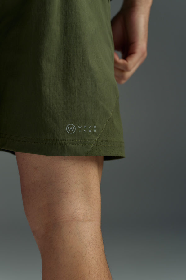 A close-up of someone wearing olive green WearEver Agile Shorts with a 7-inch inseam, showing their lower thigh and hand. A small WearEver logo appears near the hem, and the background is out of focus.