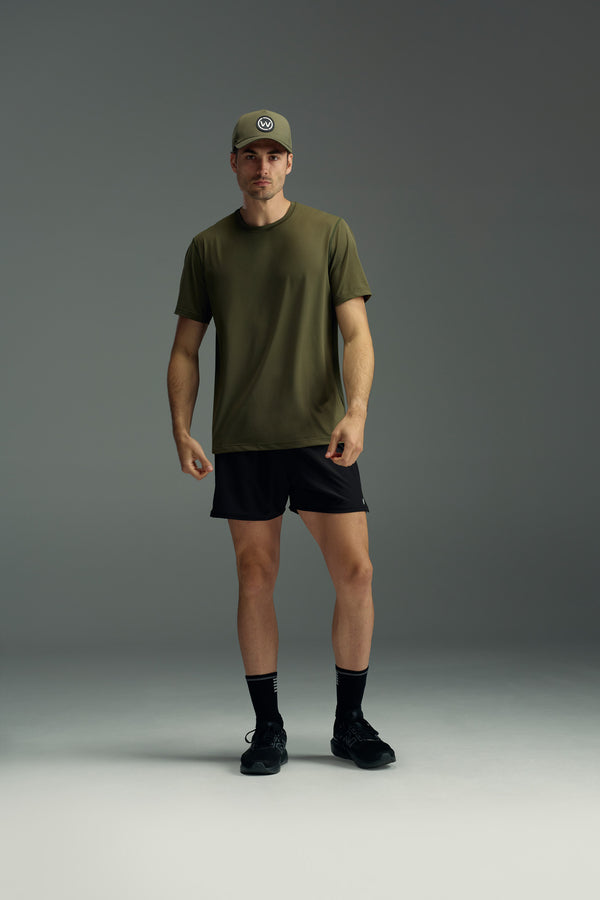 A man stands against a gray background, wearing an olive green t-shirt, the WearEver Cadence Short by WearEver in black with a 5-inch inseam, black socks, black sneakers, and an olive green cap. He looks at the camera with a neutral expression.