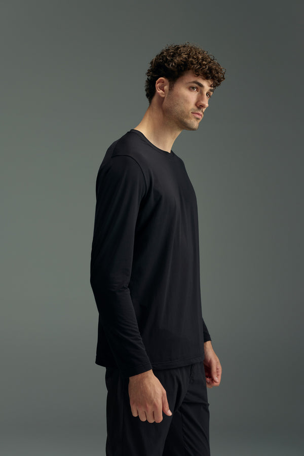 A person with short curly hair stands sideways against a plain gray background, wearing the WearEver Pulse Long Sleeve Tee by WearEver and black pants.