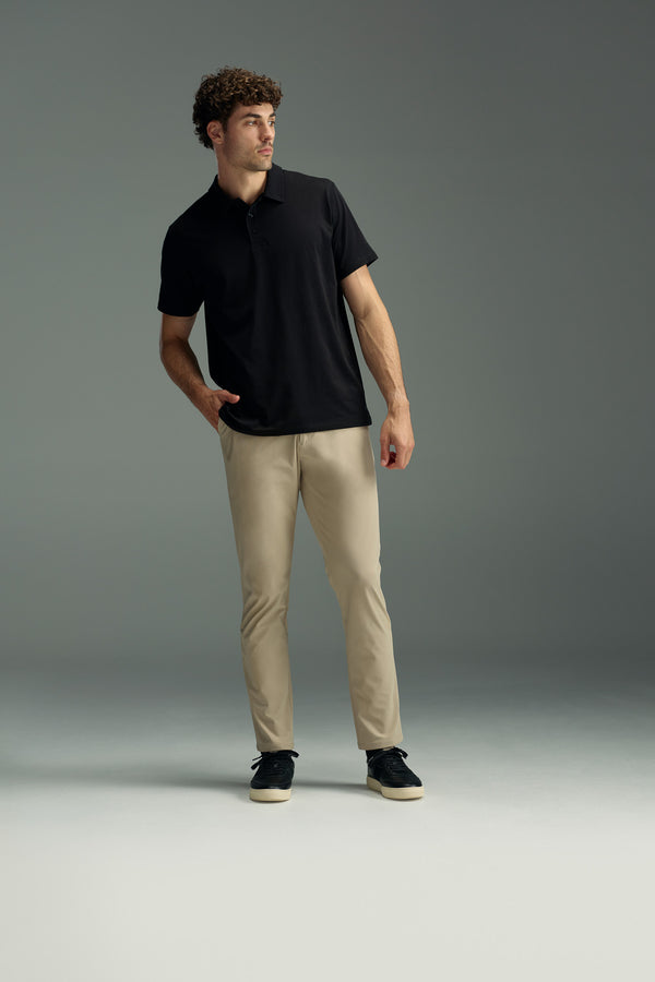 A man with curly hair stands with one hand in his pocket, looking to the side against a plain gray background. He is wearing beige WearEver Long Game Pants, a black polo shirt, and black shoes.