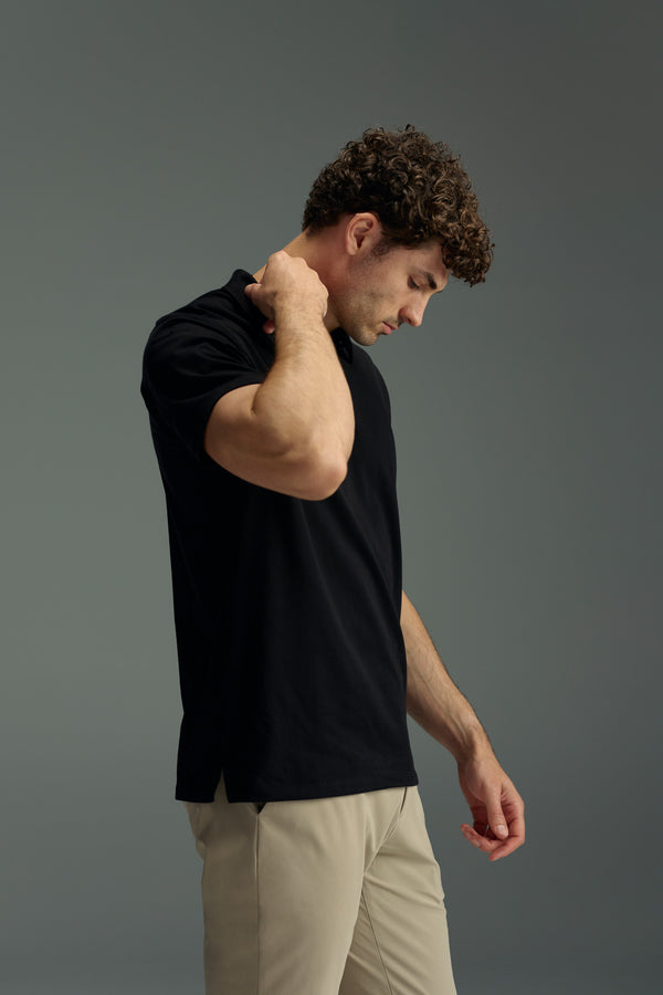 A man with curly hair wears the WearEver Outbound Polo in black and beige pants, standing against a gray background. Made from lightweight, breathable cotton-blend fabric by WearEver, this polo offers all-day comfort.
