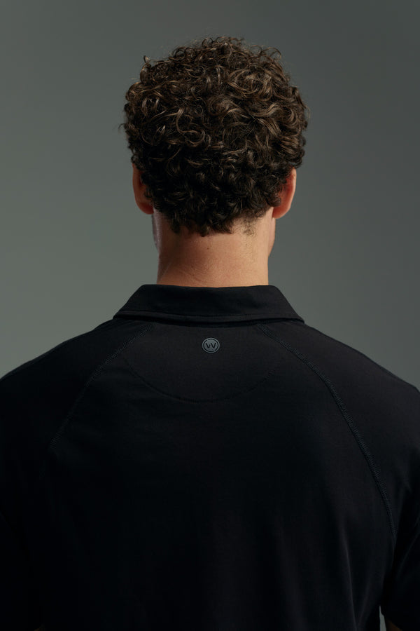 A person with short, curly brown hair is seen from behind, wearing a black WearEver Outbound Polo made from breathable cotton-blend fabric against a plain gray background.