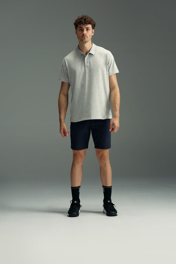 A man with short curly hair stands facing forward, wearing a light gray polo shirt, WearEver Switch Short by WearEver in navy blue, black socks, and black sneakers against a plain gray background.