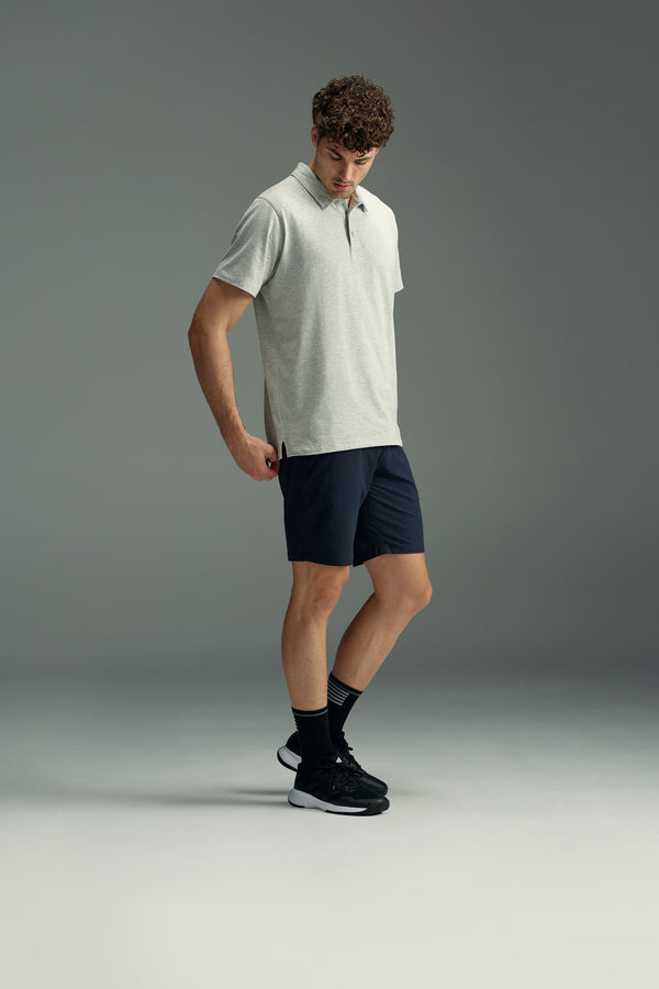 A man wearing a light grey polo, WearEver Switch Short in navy, black crew socks, and black sneakers stands looking down against a plain grey background.