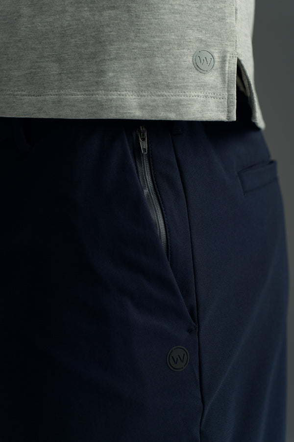 Close-up of a person wearing the WearEver Switch Short in dark navy, featuring a visible zipper pocket and the WearEver circular "W" logo. The shorts are paired with a light gray shirt with the same logo.