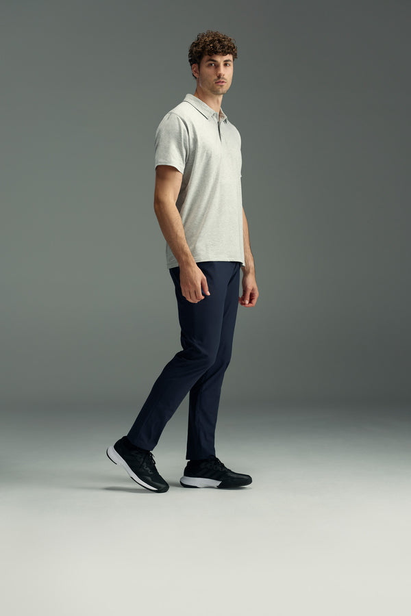 A man with curly hair wears a light gray polo shirt, WearEver Long Game Pant by WearEver in navy, and black sneakers. He stands in a neutral pose against a plain gray background.