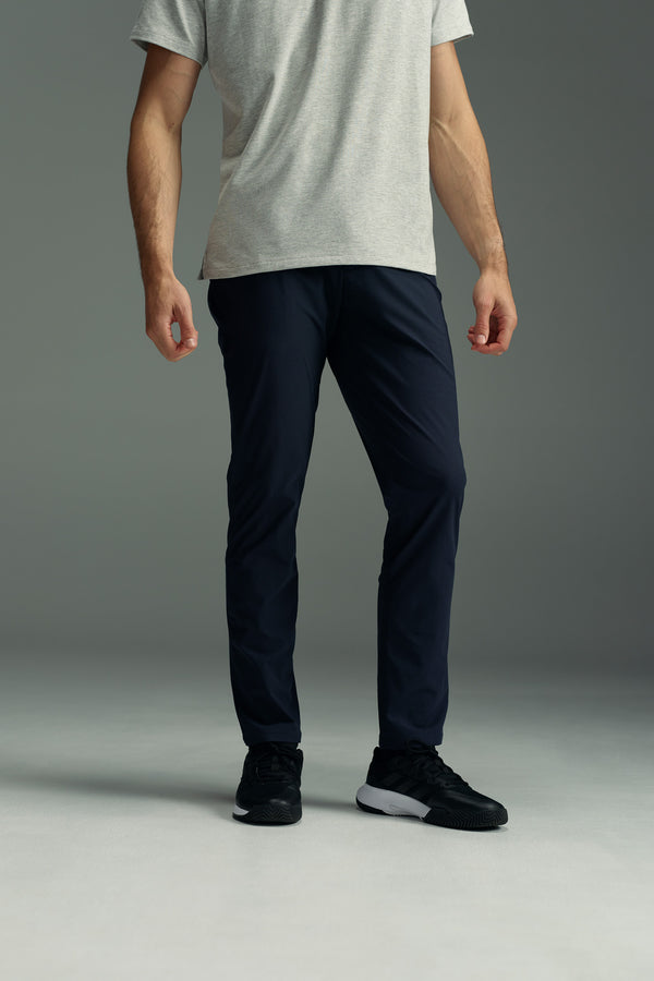 Only the lower half of a person is visible, wearing WearEver Long Game Pant by WearEver in dark navy with a chino-inspired design, paired with black sneakers and standing against a plain gray background.