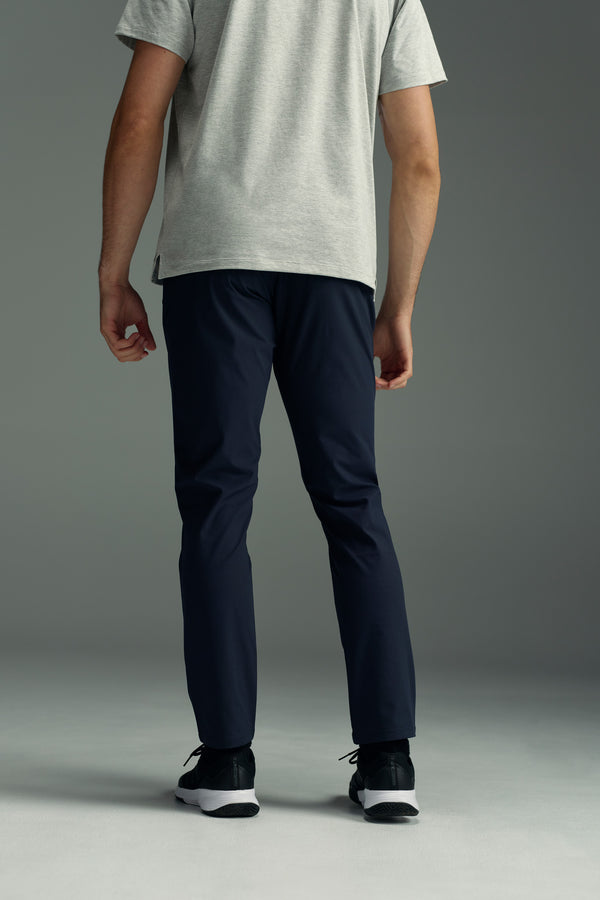 A person stands with their back to the camera, wearing a light gray t-shirt and WearEver Long Game Pant by WearEver in dark blue, featuring a chino-inspired design, along with black athletic shoes against a plain gray background.