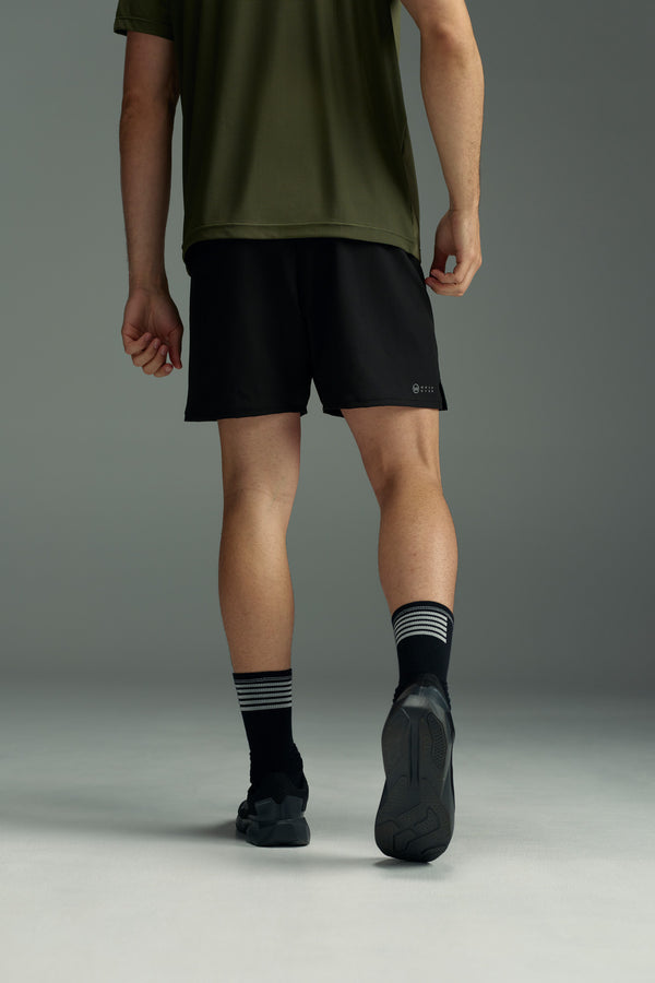 A person in a green shirt, black WearEver Cadence Short (5-inch inseam) by WearEver, black striped socks, and black sneakers is walking away from the camera against a plain gray background.