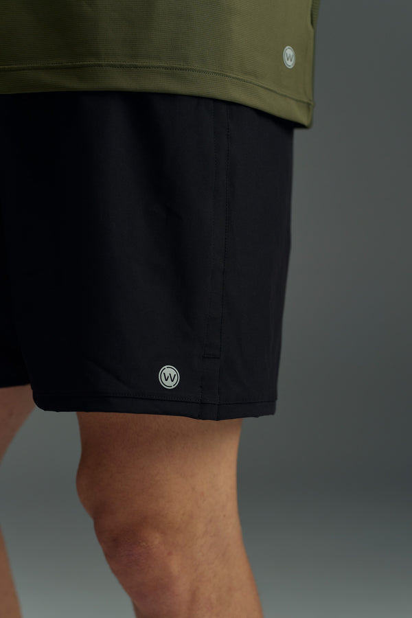 A close-up shows a person wearing the WearEver Cadence Short in black (5-inch inseam) and an olive green top, both by WearEver with small circular logos near the hem. The background is plain and neutral.
