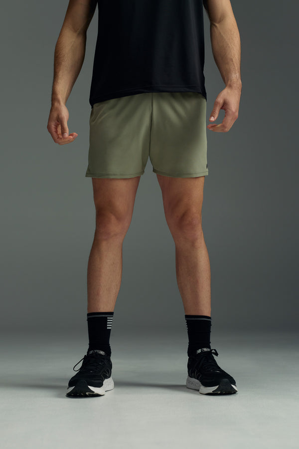 A person stands with arms relaxed at their sides, wearing a black T-shirt and WearEver Cadence Short lightweight running shorts with antibacterial treatment, paired with black socks and athletic shoes, against a plain gray background. Face not visible.