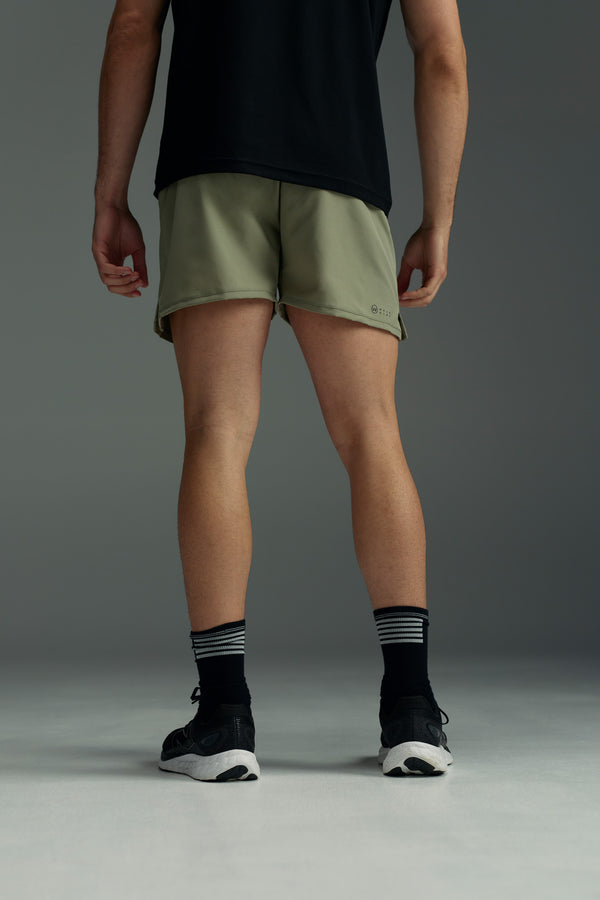 A person stands facing away, wearing a black T-shirt and olive green WearEver Cadence Short—lightweight, breathable athletic shorts with a 5-inch inseam—paired with black socks with white stripes and black sneakers against a plain gray background.
