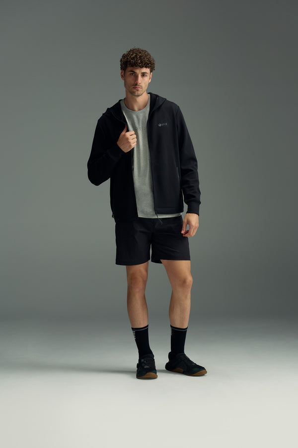 A man with curly hair stands against a gray background wearing the WearEver Altitude Hoody by WearEver, layered over a gray t-shirt with black shorts, socks, and shoes. He holds one side of his hoody collar and looks forward.