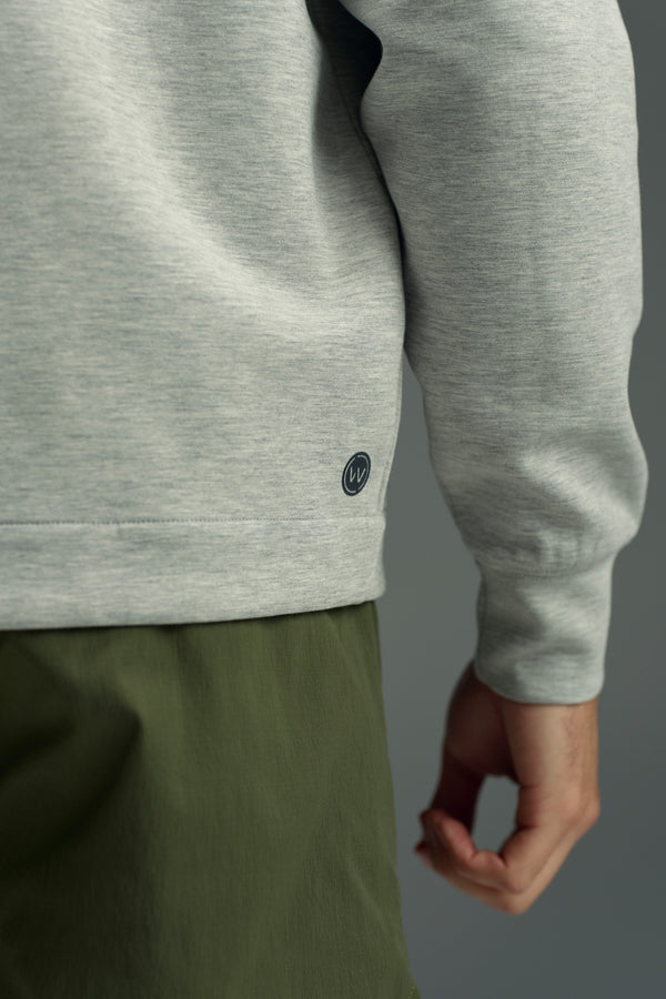 A close-up of a person wearing the WearEver Altitude Hoody in light gray, featuring a small circular logo near the hem, paired with olive green pants. Only part of the arm and hand is visible against a plain background.