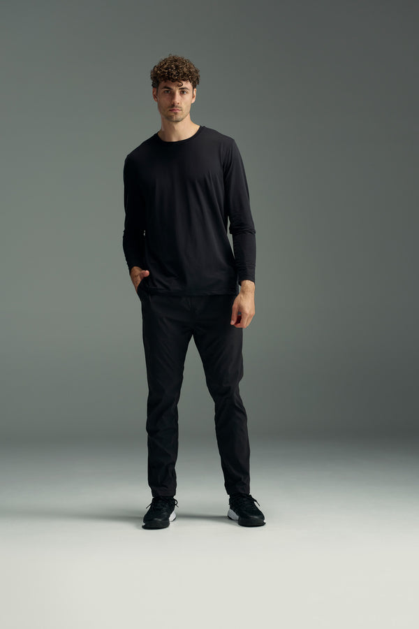 A man with curly hair stands against a gray background, wearing the WearEver Pulse Long Sleeve Tee by WearEver, paired with black pants and black sneakers, with one hand in his pocket.
