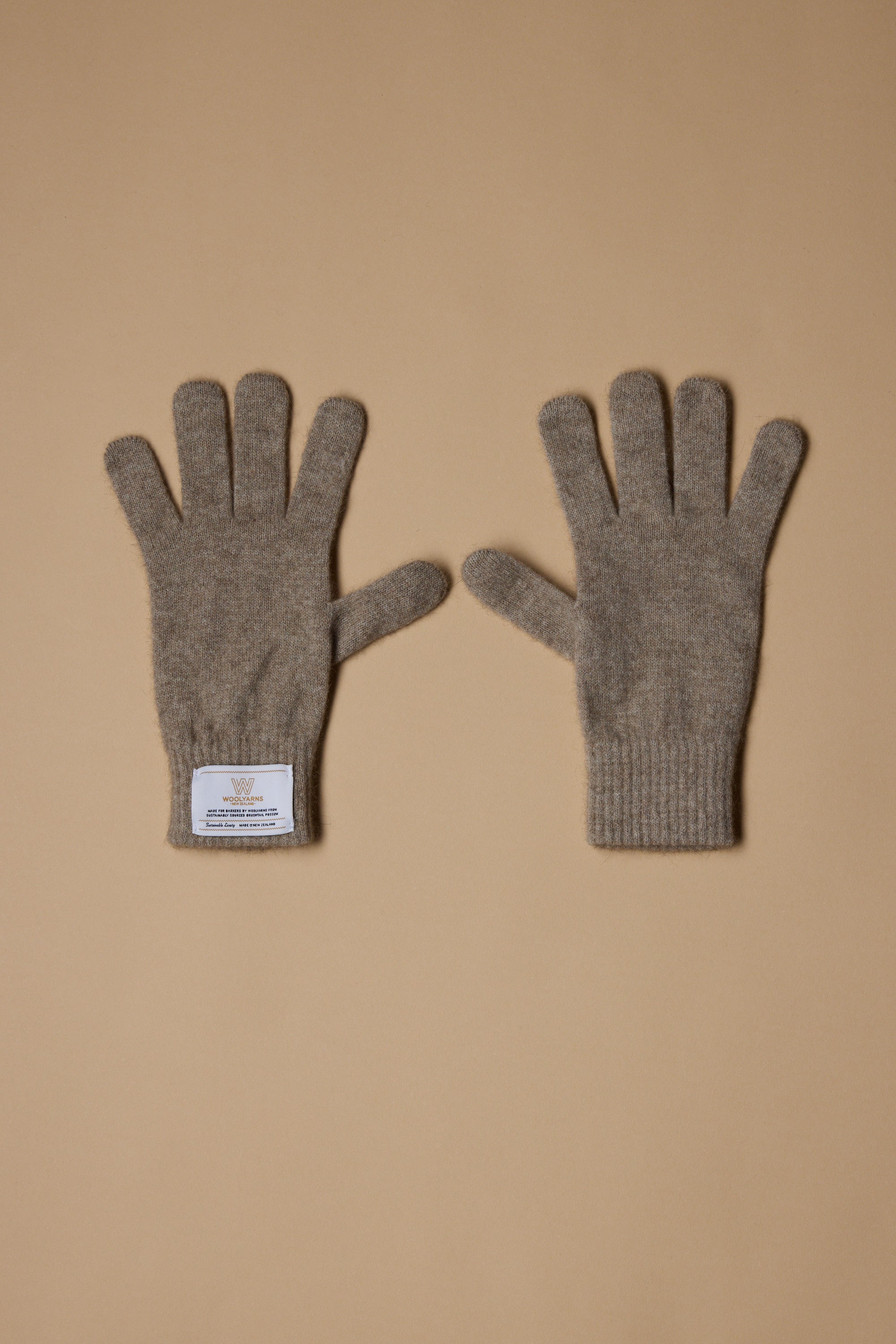 A pair of gray NZ Made Perino Gloves by Woolyarns Perino lies flat on a beige surface, crafted from luxurious merino possum silk. The left glove features a white wrist label, emphasizing these premium New Zealand accessories.
