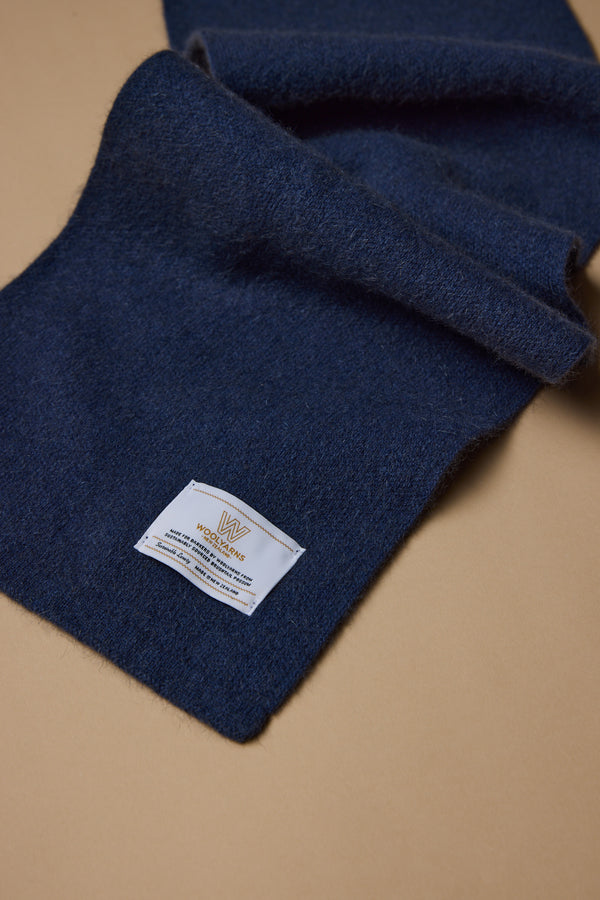 The NZ Made Perino Scarf by Woolyarns Perino is a dark blue merino possum silk blend, with a white fabric label in gold and blue text, displayed on a light brown surface. Crafted sustainably in New Zealand as part of the Perino accessories range.