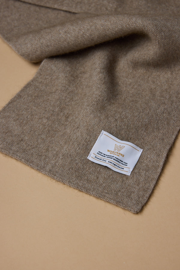 A close-up of the NZ Made Perino Scarf by Woolyarns Perino, a soft brown merino possum silk blend, laid on beige with a white label featuring text and the Woolmark logo in one corner.