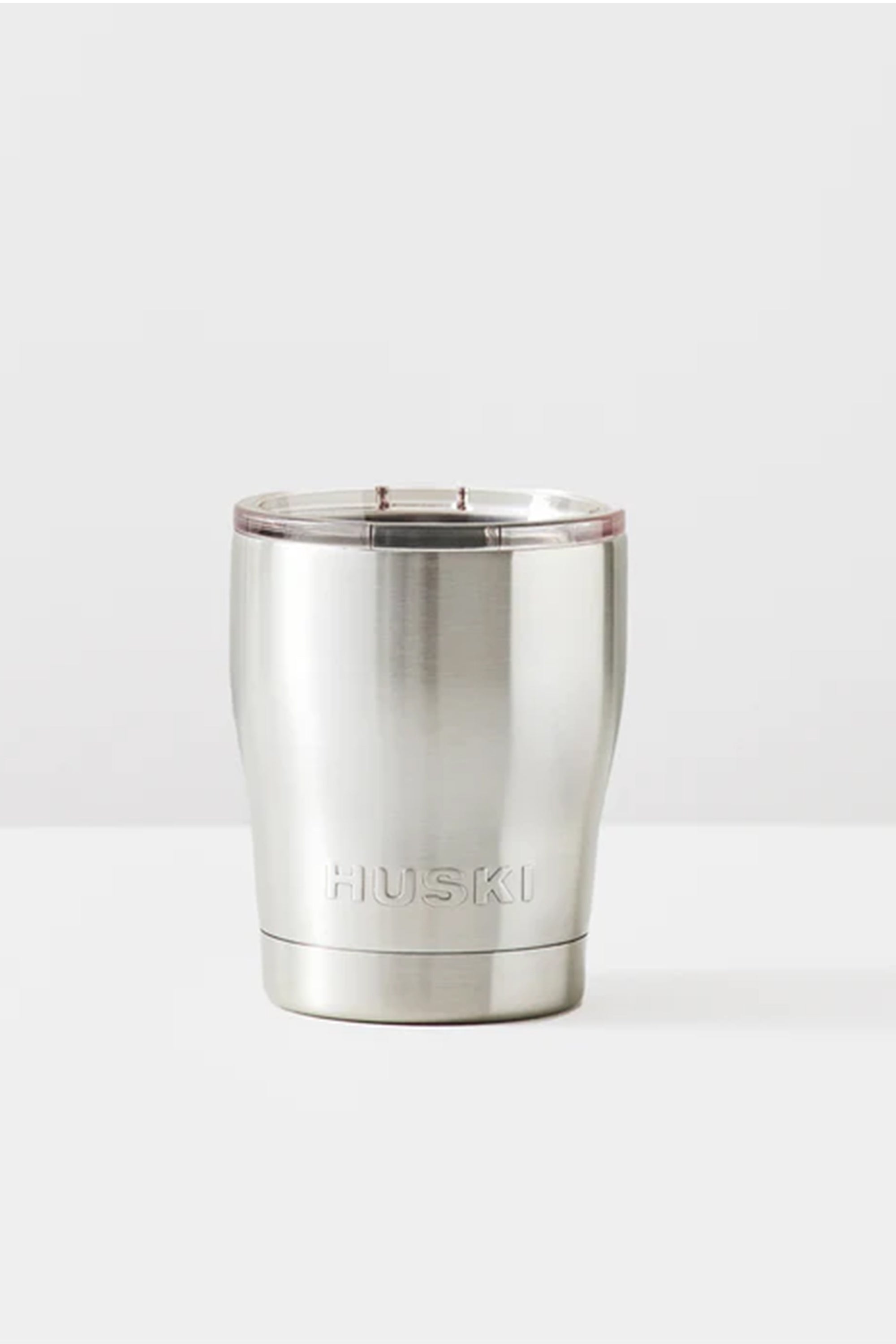 The Huski Short Tumbler 2.0 is a stainless steel, vacuum insulated tumbler with a clear plastic lid and the Huski logo engraved on the front. It sits on a white surface with a light grey background.