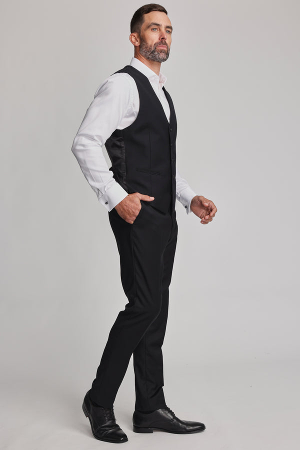 A man with short hair and a beard wears a white shirt, Barkers Mayfair ZQrx Waistcoat, black dress pants, and black dress shoes, standing in a studio with a light gray background.