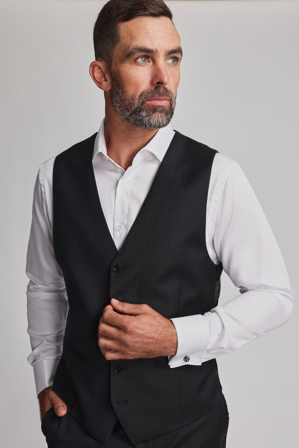 A man with short dark hair and a beard wears the Barkers Mayfair ZQrx Waistcoat over a white dress shirt, looking to the side with a serious expression. He holds one side of the vest against a plain, light background.