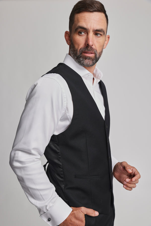A man with short dark hair and a trimmed beard wears the Barkers Mayfair ZQrx Waistcoat, a black ethical merino piece, over a white dress shirt. He stands against a plain light background, looking to the side with one hand in his pocket.