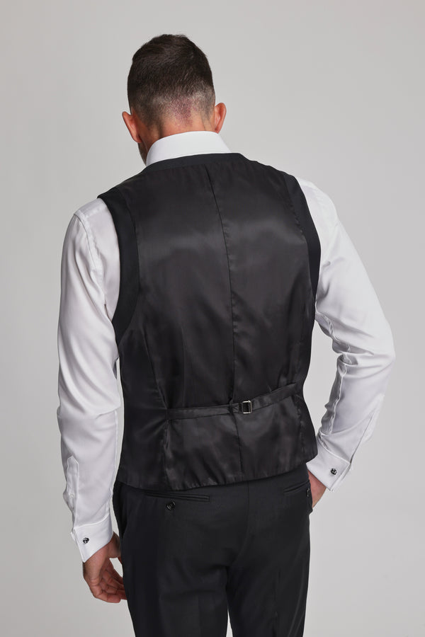 A man faces away, wearing the Barkers Mayfair ZQrx Waistcoat in black ZQrx Merino Wool with an adjustable back strap, paired with a white shirt and black trousers, set against a plain light background.