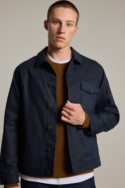 A young man with short hair wears the Barkers Benson Waxed Cotton Jacket in navy over a brown ribbed sweater and white t-shirt, standing against a plain light background and looking directly at the camera with a neutral expression.