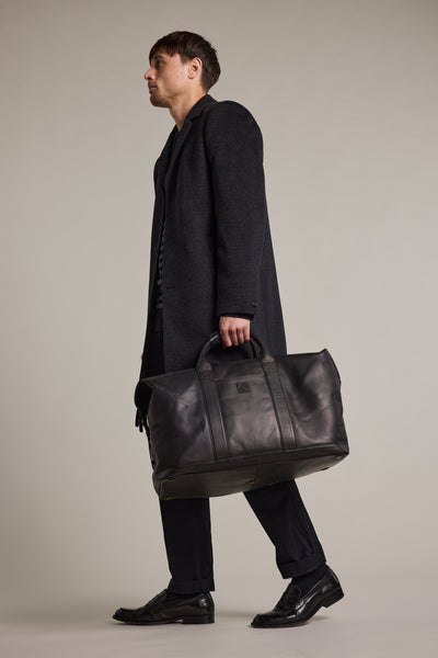 A man in a dark overcoat, black pants, and black shoes walks left against a light background, carrying the Barkers Costello Leather Weekender bag.