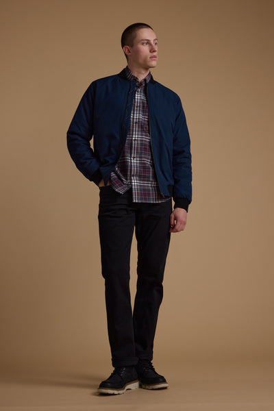 A man with short hair stands against a plain brown background, wearing the Barkers Dunhill Bomber in navy over a red plaid shirt, paired with black jeans and black shoes. One hand is in his pocket as he looks slightly to the side.