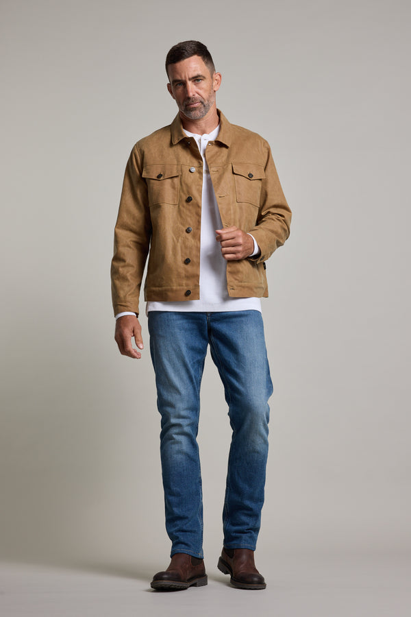 A man with short dark hair and a neutral expression stands against a plain background, wearing the Barkers Benson Waxed Cotton Jacket in tan over a white shirt, blue jeans, and brown boots.