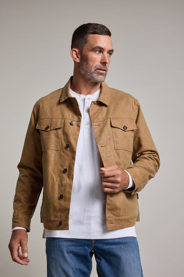 A man with short brown hair and a trimmed beard wears a tan Barkers Benson Waxed Cotton Jacket over a white shirt and blue jeans, standing against a plain light gray background and looking to the side with a neutral expression.