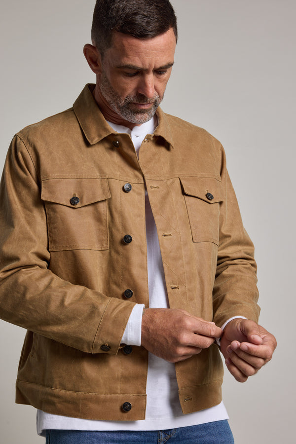 A man with short dark hair and a beard wears a Barkers Benson Waxed Cotton Jacket in tan over a white shirt and blue jeans, looking down as he fastens the cuff. The background is plain and light-colored.