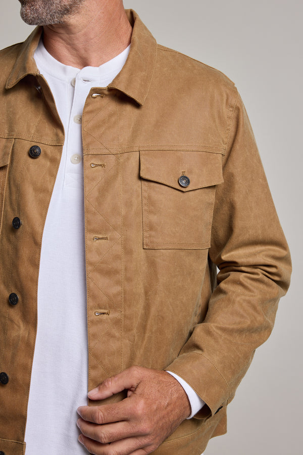 A man wears the Barkers Benson Waxed Cotton Jacket in light brown over a white henley shirt, shown from shoulders to waist, holding the jacket's edge with one hand.