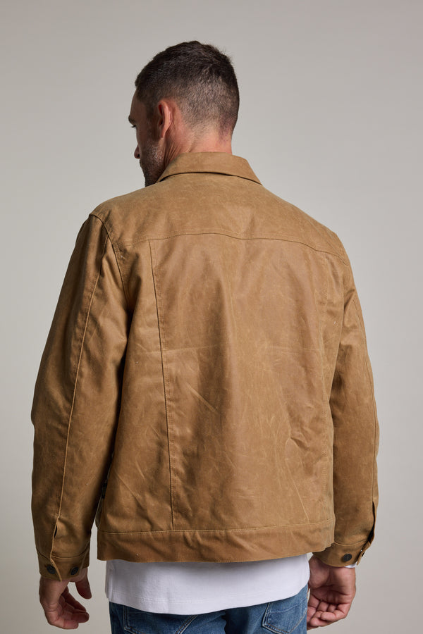 A man stands with his back to the camera, wearing a tan Barkers Benson Waxed Cotton Jacket over a white shirt and blue jeans, set against a plain light background.