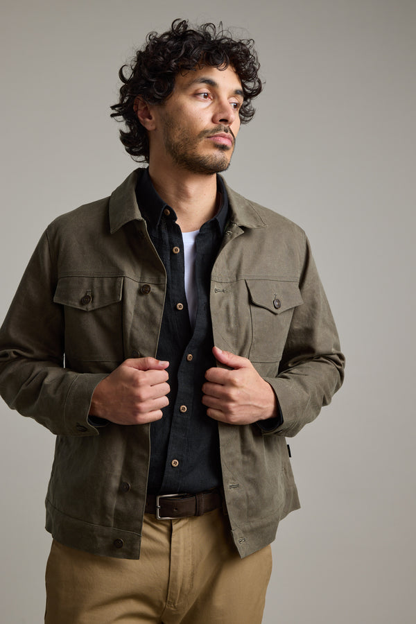 A man with curly hair wears the Barkers Benson Waxed Cotton Jacket over a black shirt and tan pants, standing against a plain light gray background. He looks slightly to the side with his hands holding the jacket.