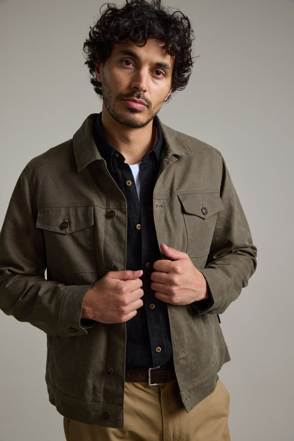 A man with curly hair and a short beard wears the Barkers Benson Waxed Cotton Jacket in olive green over a black shirt and khaki pants, standing against a plain light background and looking calmly at the camera.